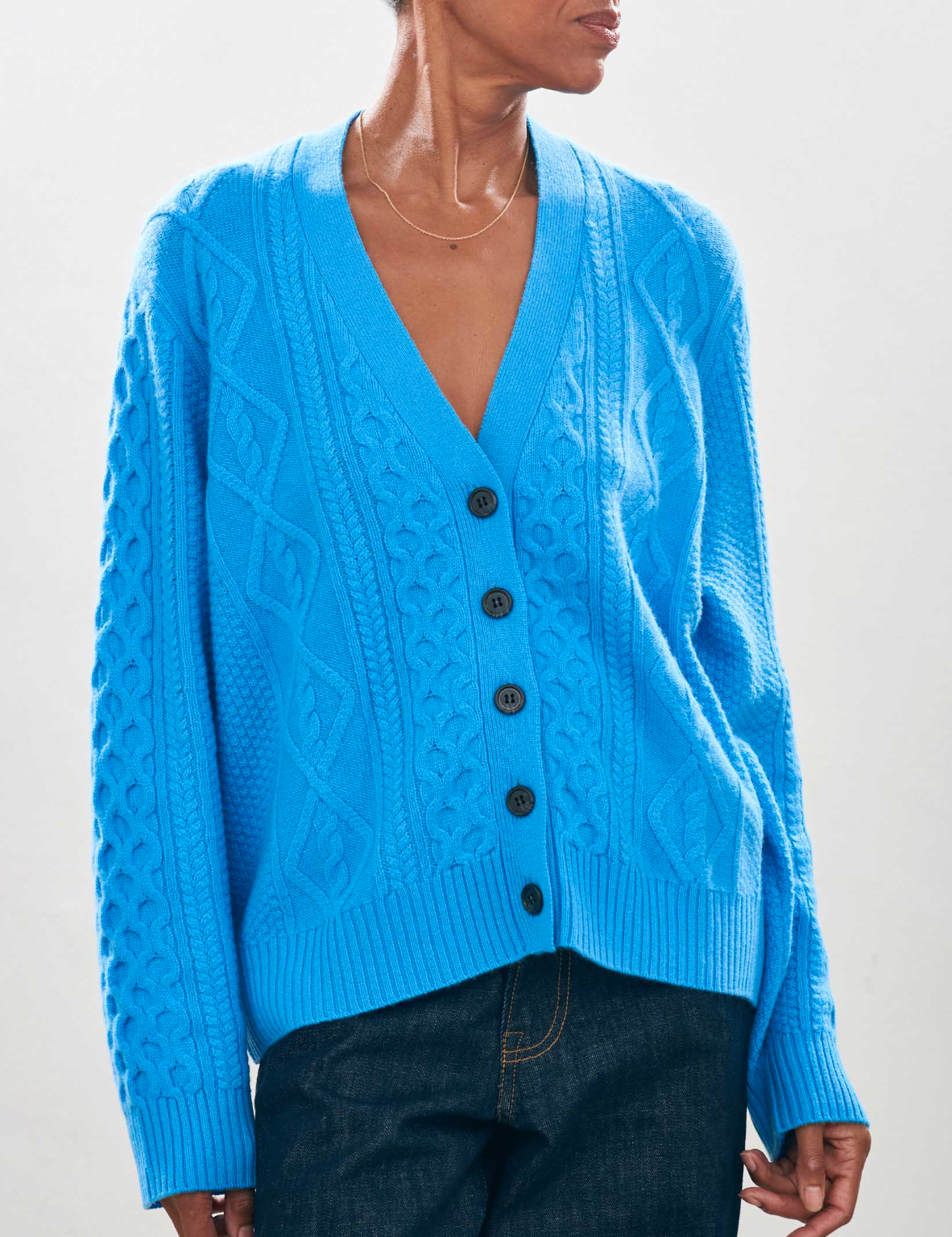 A person is wearing an Aran Cardigan from Jumper 1 2 3 4/CR2, featuring a bright blue, oversized design made from cashmere wool with a V-neck and black buttons. This cable-knit piece pairs seamlessly with dark jeans against a plain background.