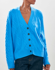 A person is wearing an Aran Cardigan from Jumper 1 2 3 4/CR2, featuring a bright blue, oversized design made from cashmere wool with a V-neck and black buttons. This cable-knit piece pairs seamlessly with dark jeans against a plain background.