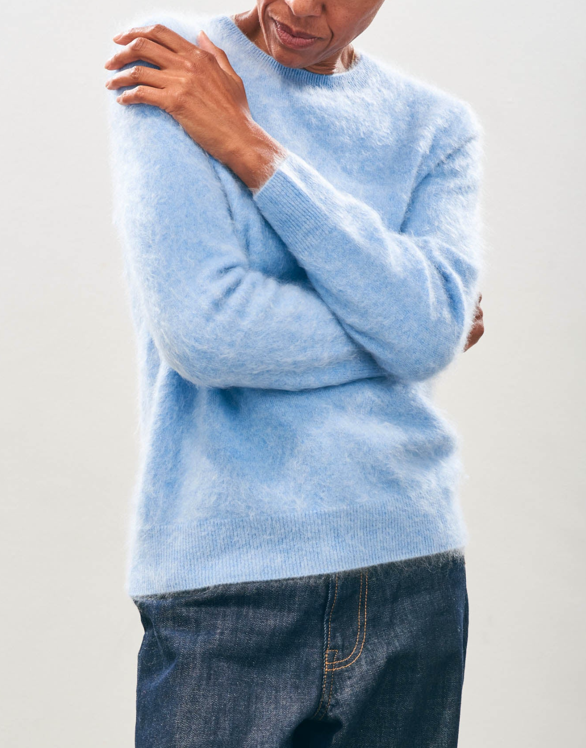 A person wearing a Brushed Crew sweater from Jumper 1 2 3 4/CR2 in a soft blue shade and paired with dark denim jeans, standing with arms crossed against a plain background.