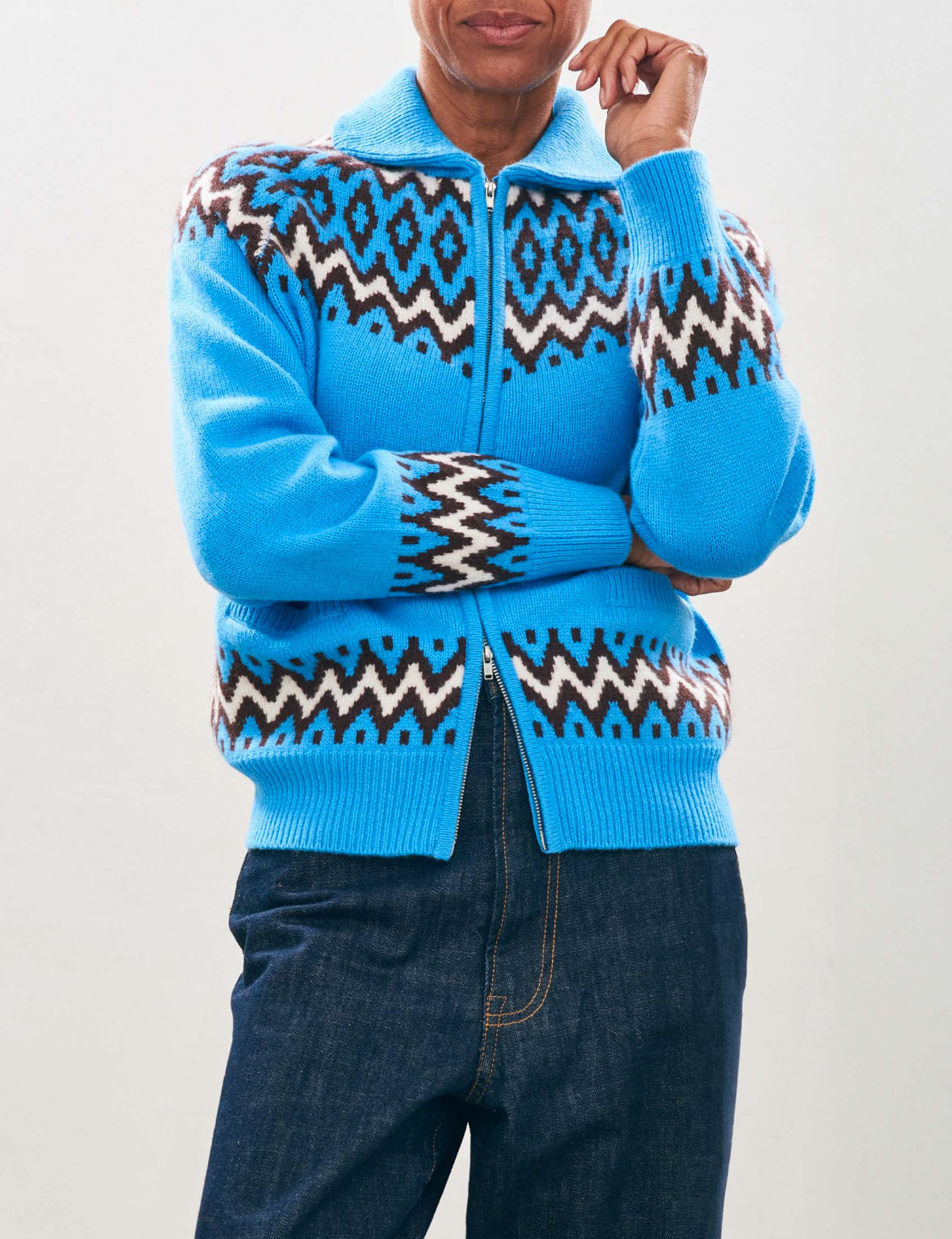 A person is wearing the Jumper 1 2 3 4/CR2 Ski Zipup, which is a bright blue zip-up cardigan featuring geometric patterns in white, brown, and black that resemble a zig zag jacquard design. It pairs perfectly with dark blue jeans against a simple light background.