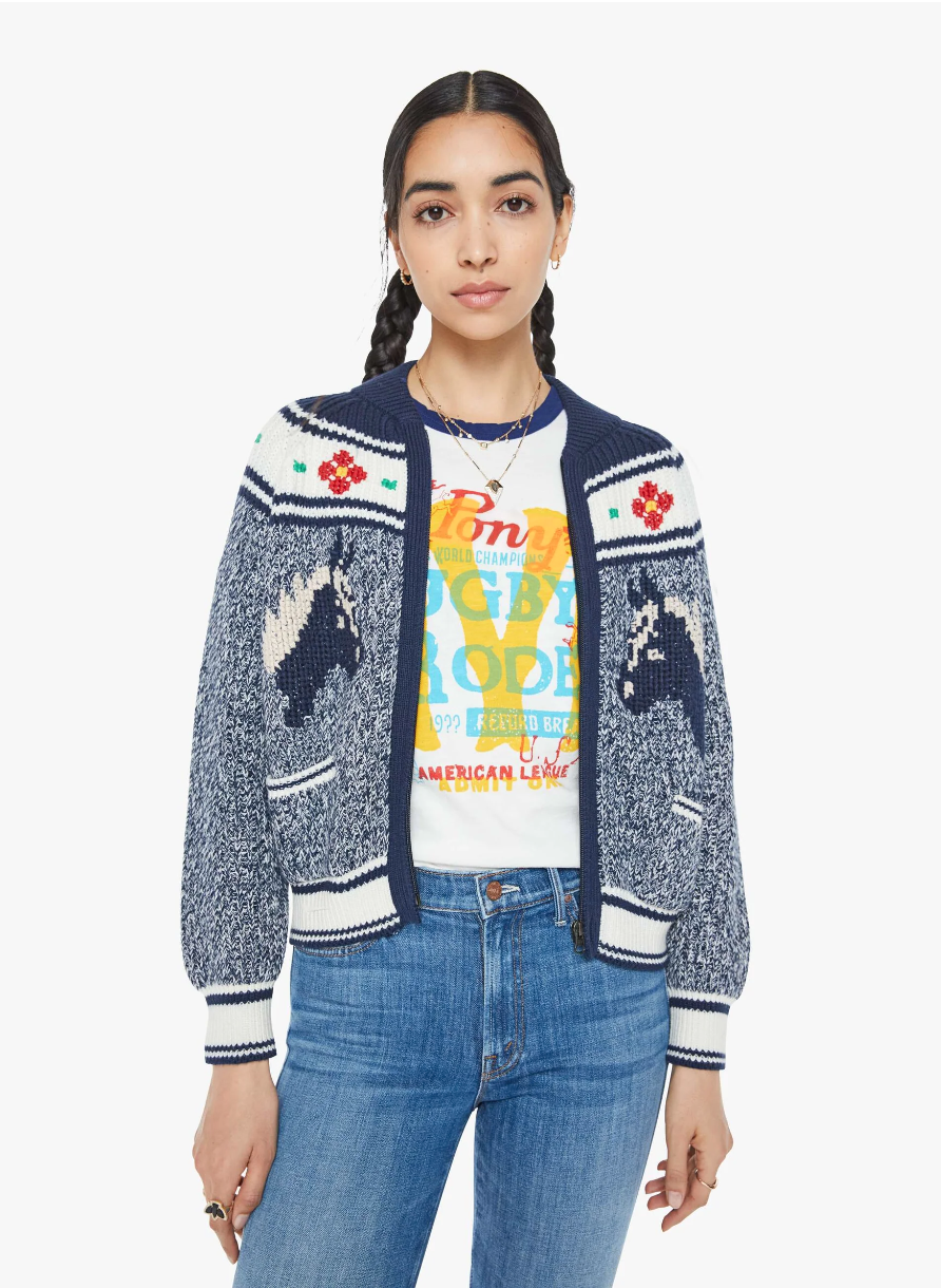 A person with long dark hair poses in "The Try Outs" crewneck cardigan sweater by Mother, featuring marled navy and white horse designs, paired with a colorful graphic T-shirt and blue jeans. They have a neutral expression against a plain white background.