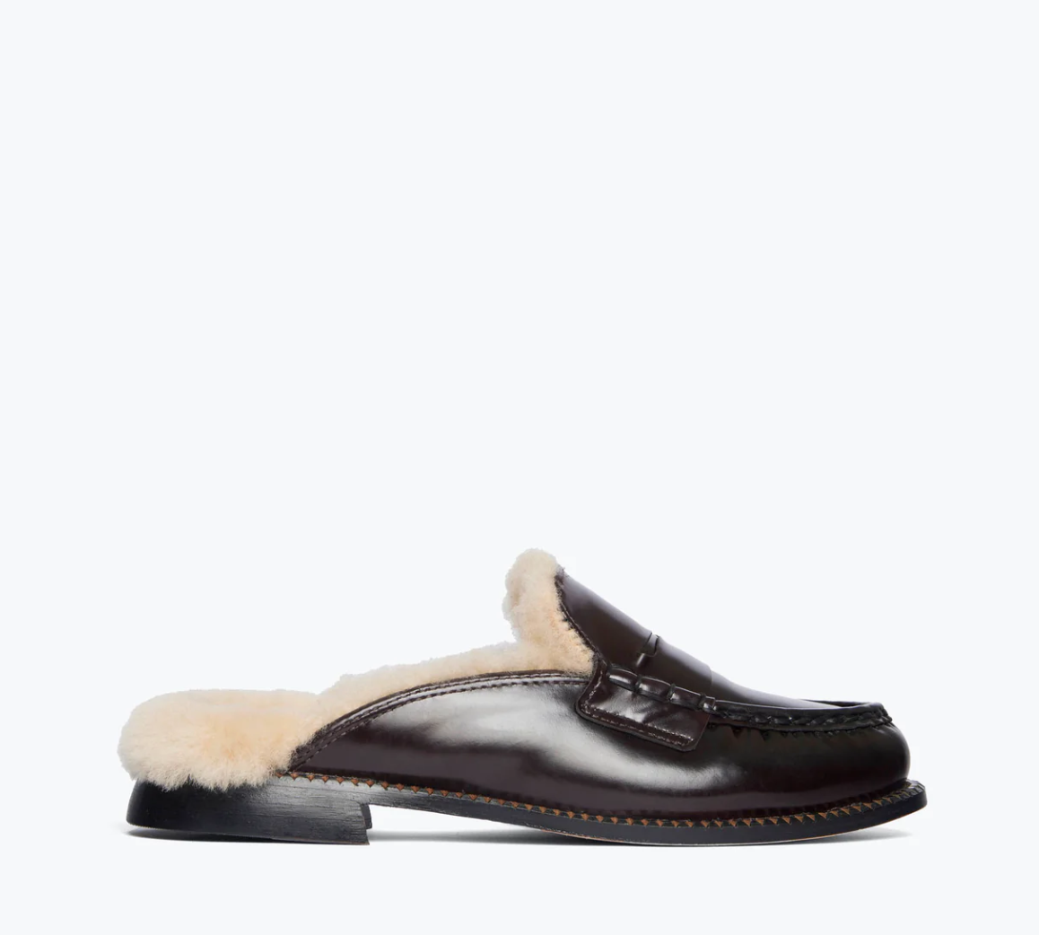 The TEMI SHEARLING PENNY LOAFER MULE by Freda Salvador is a brown leather shoe crafted from high-quality Italian leather, featuring a luxurious, cream-colored shearling lining. It showcases a smooth, polished finish with a classic loafer design enhanced by intricate stitching details. The ethically sourced shearling lining is visible around the edges.