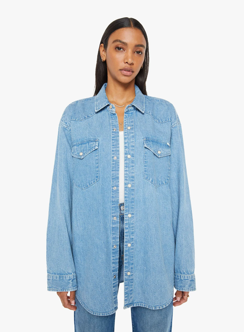 A person stands against a plain white background wearing "The Exes And The Ohs," an oversized denim button-up in a vintage-blue wash by Mother. The shirt is detailed with button-down pockets and faux pearl snaps. They have long, dark hair and a neutral expression.