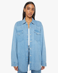 A person stands against a plain white background wearing "The Exes And The Ohs," an oversized denim button-up in a vintage-blue wash by Mother. The shirt is detailed with button-down pockets and faux pearl snaps. They have long, dark hair and a neutral expression.