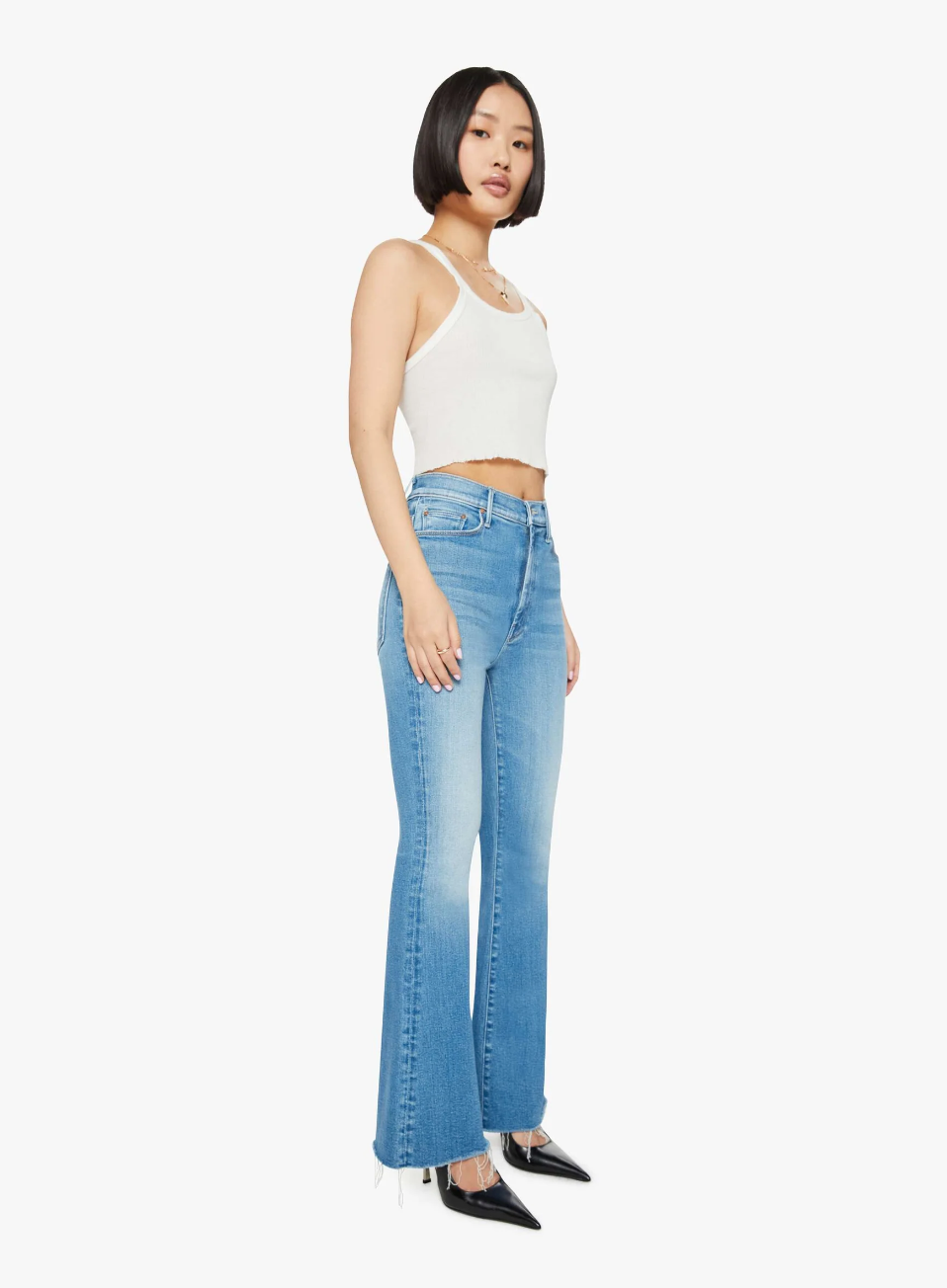 A person with a bob haircut is wearing a white cropped tank top and the Lil HW Weekender Fray jeans by Mother, crafted from soft stretch denim. They stand slightly to the side, hands by their sides, against a plain white backdrop.