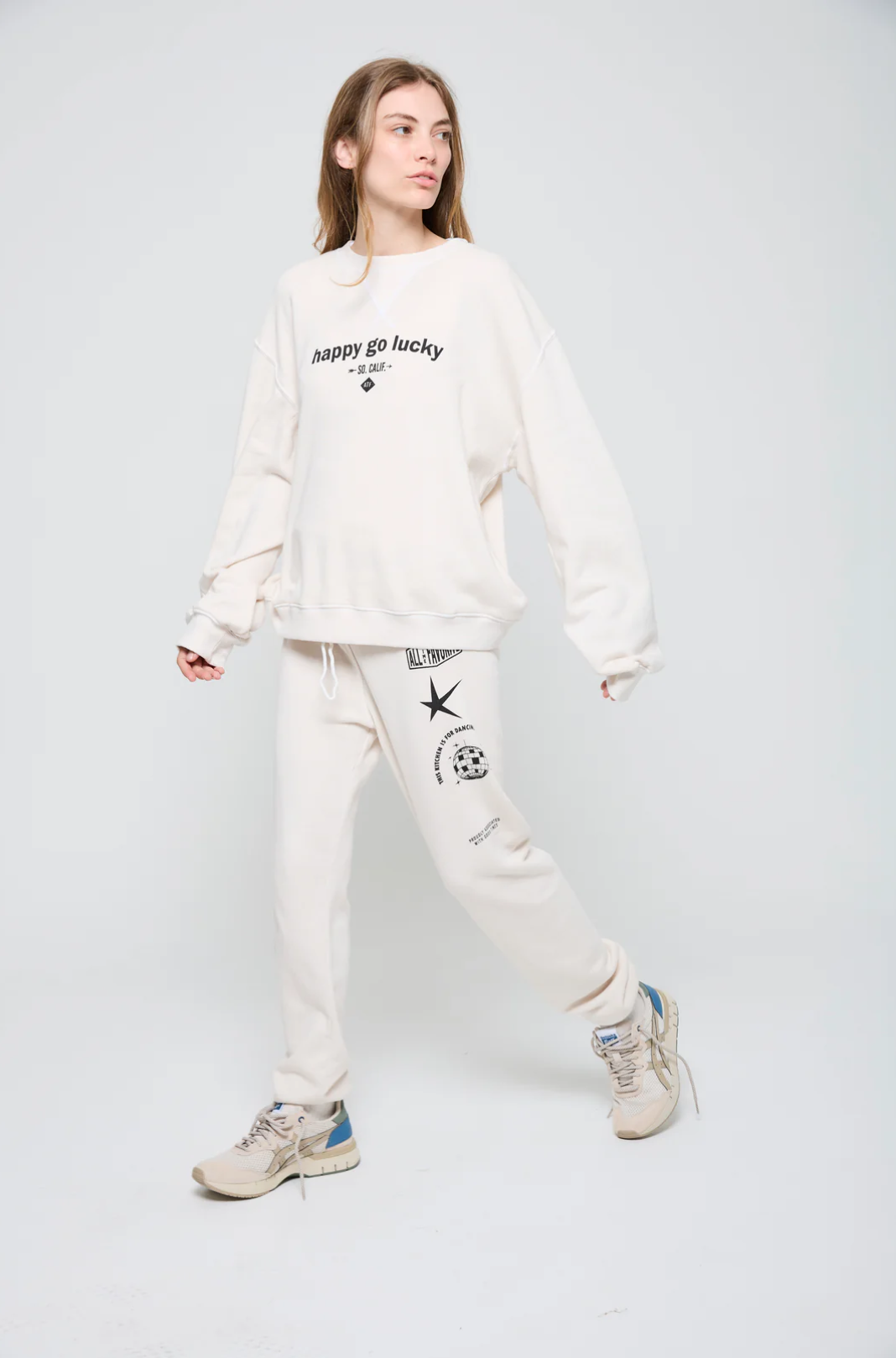 A person confidently walks against a plain background, wearing an oversized fit Happy Go Lucky BF Sweatshirt from ATF Clothing. Their cozy brushed terry top showcases the "happy go lucky" text, paired with matching sweatpants featuring black designs. They complete the look with beige sneakers accented in blue.