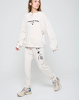 A person confidently walks against a plain background, wearing an oversized fit Happy Go Lucky BF Sweatshirt from ATF Clothing. Their cozy brushed terry top showcases the "happy go lucky" text, paired with matching sweatpants featuring black designs. They complete the look with beige sneakers accented in blue.