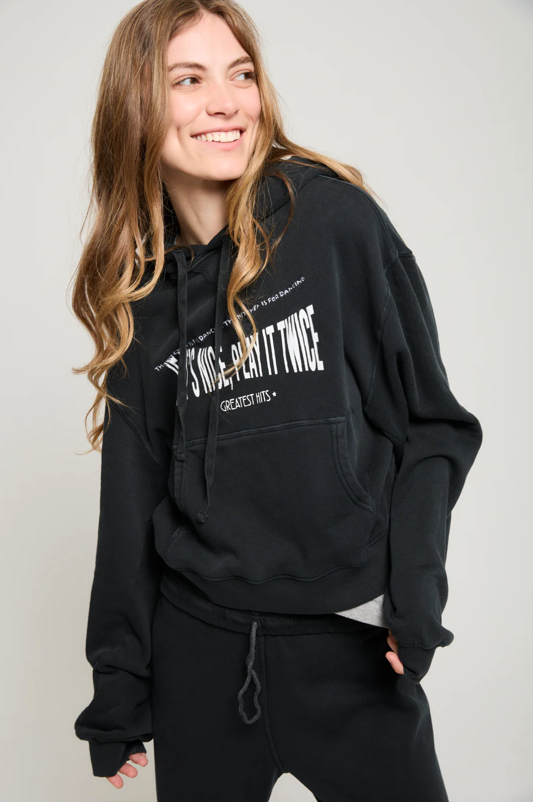 A person with long hair is smiling and looking to the side while wearing an ultra-soft black "Play it Twice Cropped Hoodie" from ATF Clothing, featuring a graphic print. They stand against a plain background, with their hands in the hoodie’s pocket and dark pants.