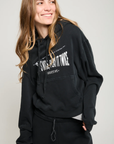 A person with long hair is smiling and looking to the side while wearing an ultra-soft black "Play it Twice Cropped Hoodie" from ATF Clothing, featuring a graphic print. They stand against a plain background, with their hands in the hoodie’s pocket and dark pants.