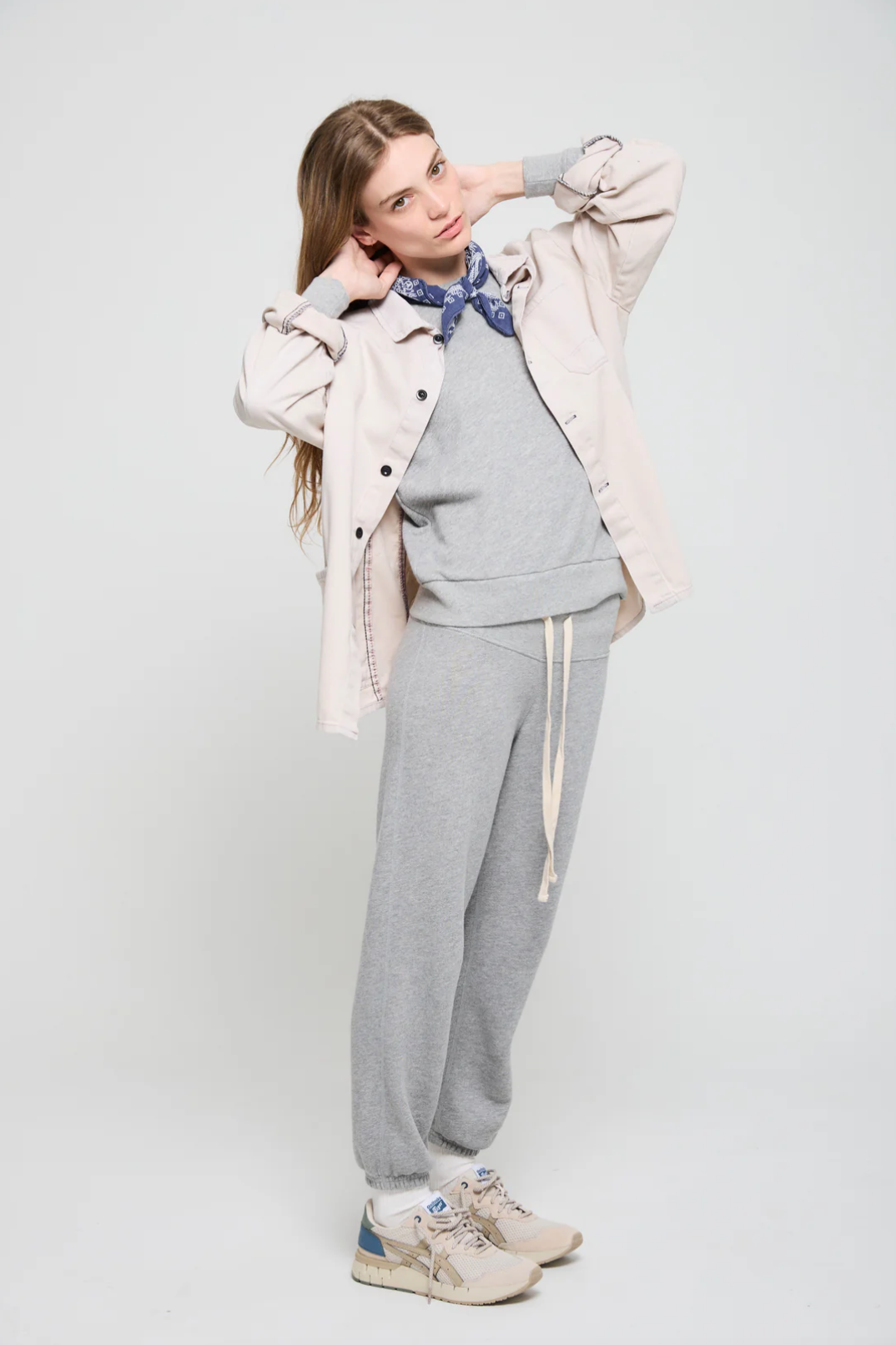 Against a light background, a person models in a beige jacket and the Venice Sweats from ATF Clothing. Beige sneakers complement the look, while a navy blue bandana adds flair as they adjust their hairstyle.