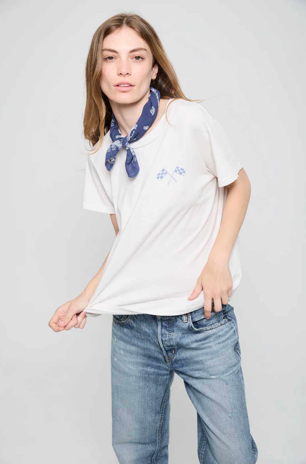 A person with long hair wears a Going Nowhere Fast SS Crew by ATF Clothing, featuring racing flag prints. They pair it with light blue jeans and accessorize with a blue bandana stylishly tied around their neck, which contrasts against the plain white shirt.