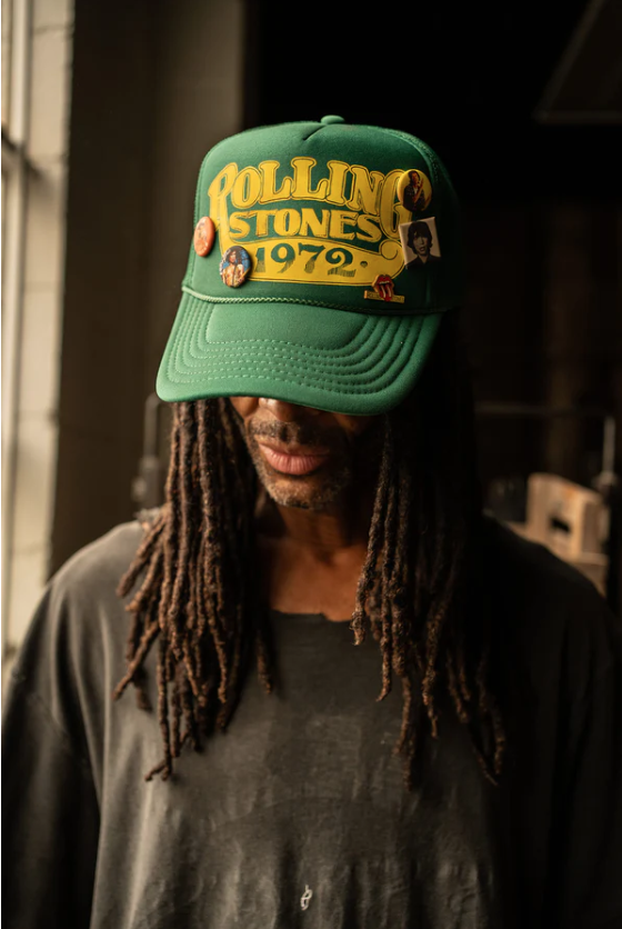 A person with long dreadlocks wears the Rolling Stones 1972 Hat from Made Worn, adorned with custom pins. The green cap, featuring an adjustable back strap for a comfortable one-size-fits-most fit, completes their look as they stand indoors. Warm lighting enhances the relaxed atmosphere.