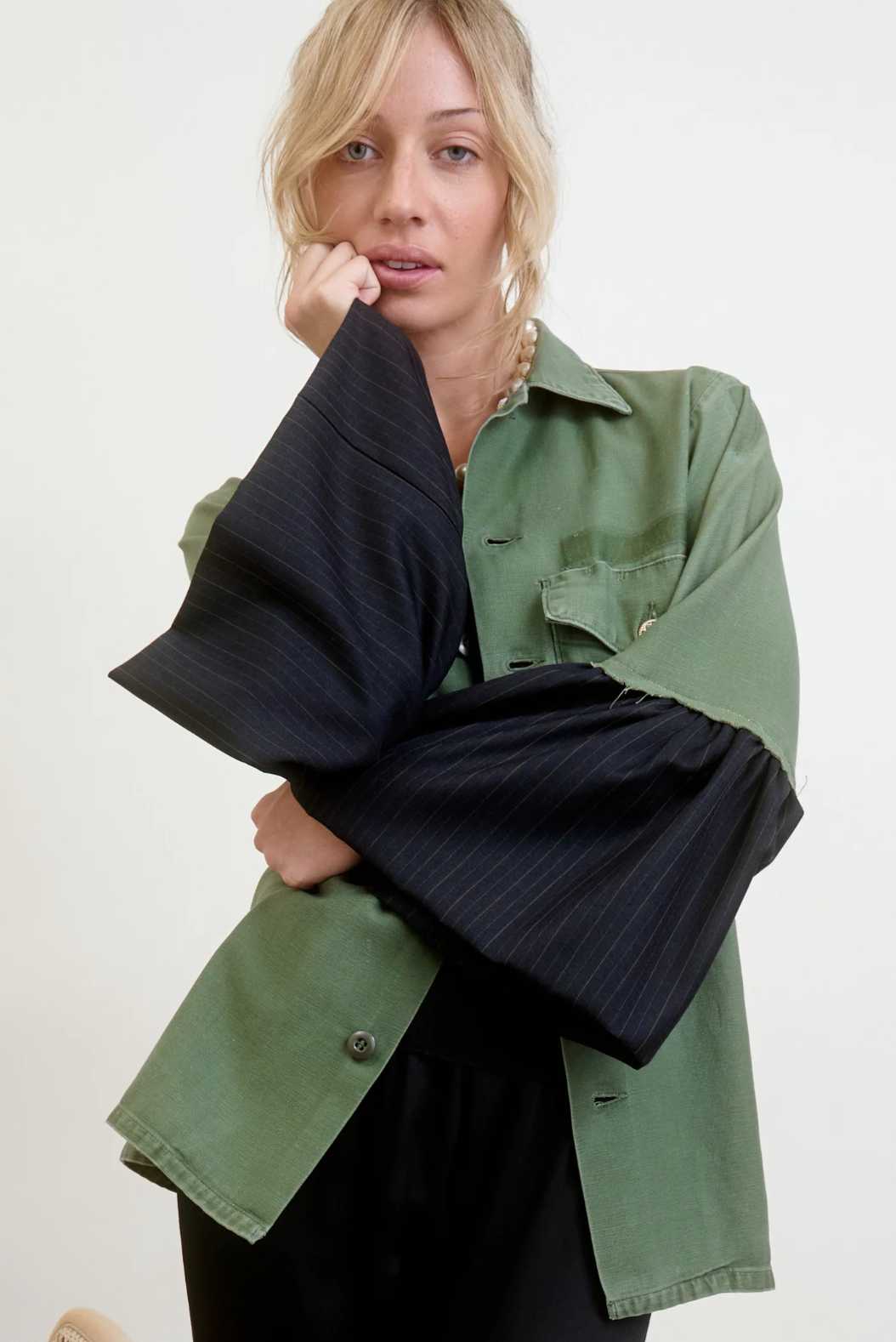 Against a plain white background, a person with blonde hair poses thoughtfully in an Aquarius Cocktail Hooper Suiting Cuff Army Jacket. This green vintage cotton army jacket, adorned with dead stock couture designer buttons and oversized black sleeves, adds a unique touch as they rest their chin on their hand.