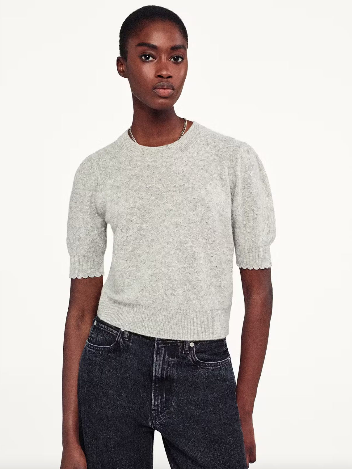 A person in a FRAME Pointelle Puff Sleeve sweater, light gray and knit with puff half sleeves, pairs it with dark jeans while standing against a plain white backdrop, looking at the camera.