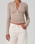 A person in a beige VARRA HENLEY ribbed organic cotton top by Citizens Of Humanity/AGOLDE and white high-waisted jeans stands against a plain background, with their right hand in their pocket and wearing a simple necklace.