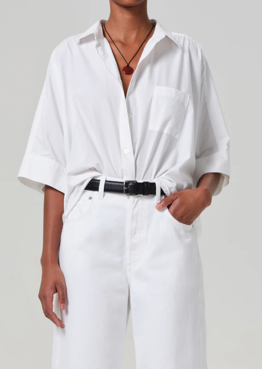 Dressed in a Claire Origami Shirt by Citizens Of Humanity/AGOLDE, with oversized sleeves tucked into white trousers and secured by a black belt. They sport a necklace with a round, dark pendant and casually place one hand in their pocket against a neutral gray background.