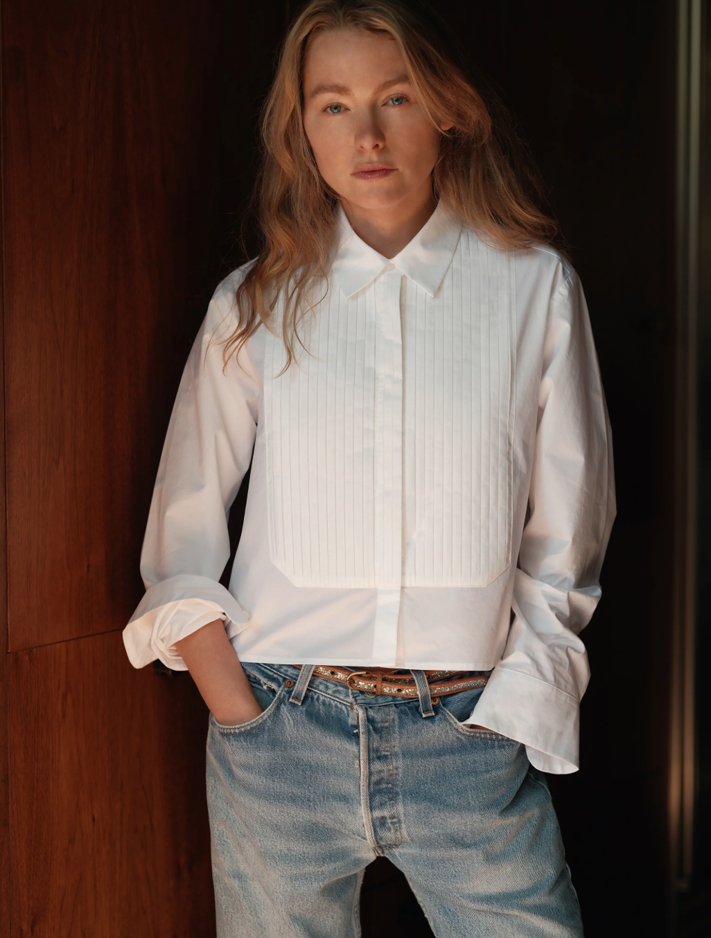 A woman with long hair, wearing Cissa's crisp The Tuxedo Shirt and blue jeans, stands confidently with her hands in pockets, leaning against a wooden surface.