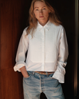 A woman with long hair, wearing Cissa's crisp The Tuxedo Shirt and blue jeans, stands confidently with her hands in pockets, leaning against a wooden surface.