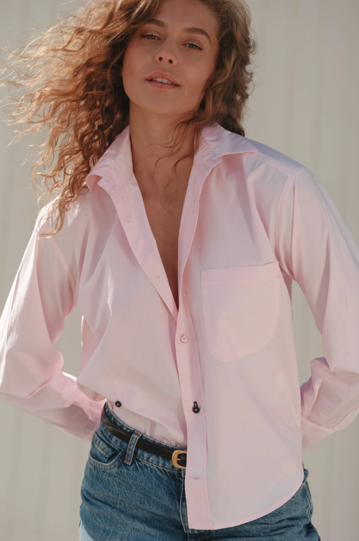 Someone with curly hair wears The Daily Shirt by Cissa, a light pink, relaxed-fit top made from PAPER COTTON, paired with blue jeans. They stand confidently with hands in pockets, looking at the camera. The softly lit background enhances the neutral tone.