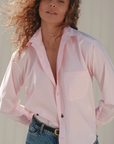 Someone with curly hair wears The Daily Shirt by Cissa, a light pink, relaxed-fit top made from PAPER COTTON, paired with blue jeans. They stand confidently with hands in pockets, looking at the camera. The softly lit background enhances the neutral tone.