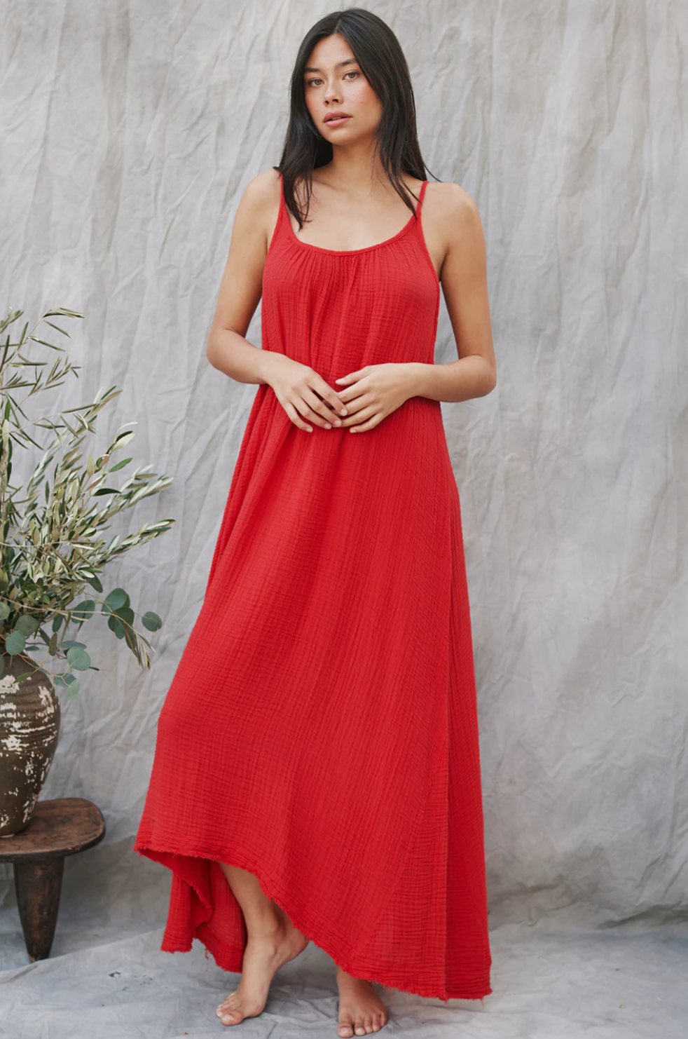 A person in a flowing red Tulum maxi dress by 9seed stands barefoot against a neutral backdrop. With long hair, they gaze forward, their arms resting gently in front. The lightweight gauze drapes elegantly, and an olive branch in a pot is visible nearby.