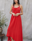 A person in a flowing red Tulum maxi dress by 9seed stands barefoot against a neutral backdrop. With long hair, they gaze forward, their arms resting gently in front. The lightweight gauze drapes elegantly, and an olive branch in a pot is visible nearby.