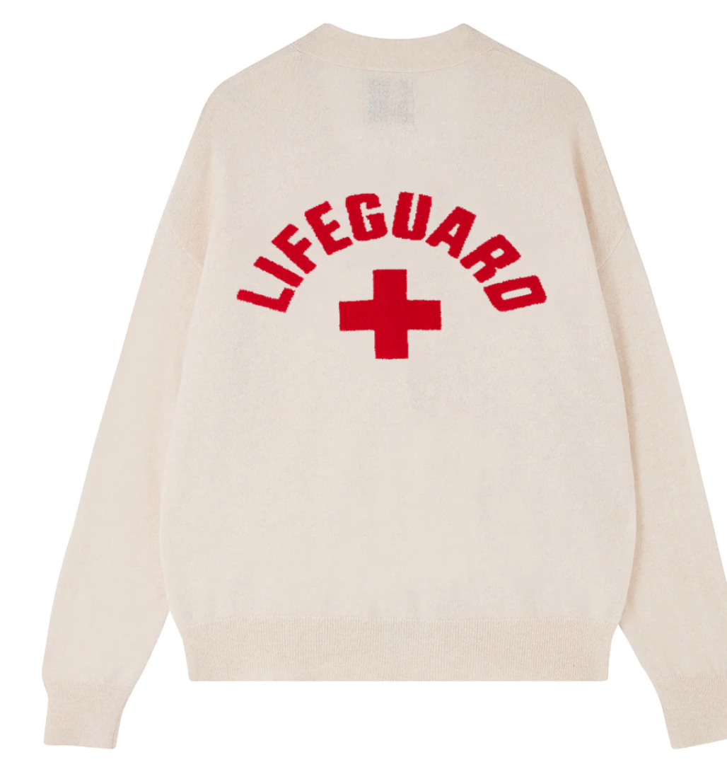 The Beach Cardigan by Jumper 1 2 3 4/CR2 is a beige cashmere piece with a red cross and "LIFEGUARD" intarsia on the back, offering both coziness and style.