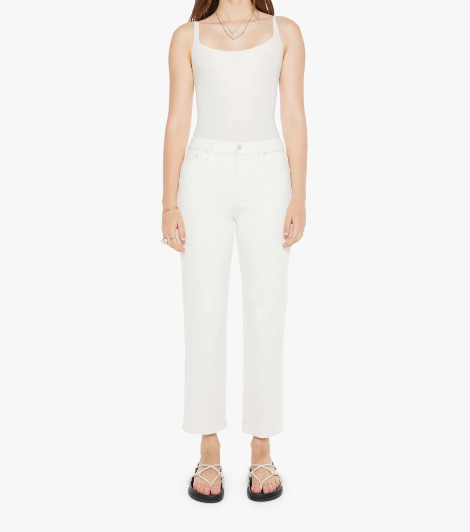 A person wearing a sleeveless white top and Mother's "The Rambler Zip Ankle" high-waisted stretch jeans stands against a plain white background. They have long hair, black sandals, and are accessorized with a few bracelets and necklaces.