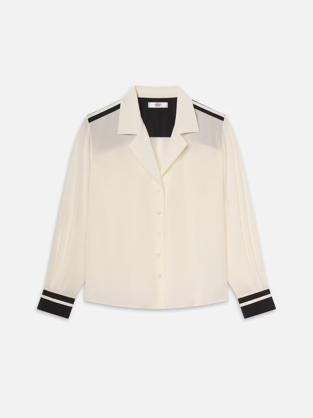 The FRAME Silk Pajama Shirt is a cream blouse with long sleeves, a button-up front, and black accents on cuffs, shoulders, and a stripe on the upper sleeve. Made from 100% silk, it features a notched collar and an elegant silhouette for a loose fit.