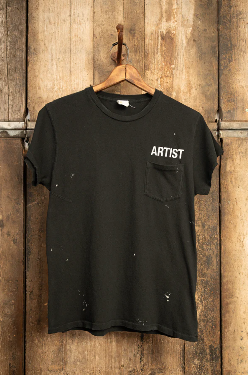 The MADE WORN "ARTIST" CROP POCKET TEE by Made Worn, is a black t-shirt featuring "ARTIST" in white on the upper left chest. It has unique distressing with paint splatters and hangs on a wooden hanger against a rustic background. This vintage unisex fit piece is handmade in Los Angeles.