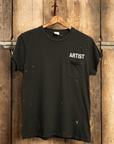 The MADE WORN "ARTIST" CROP POCKET TEE by Made Worn, is a black t-shirt featuring "ARTIST" in white on the upper left chest. It has unique distressing with paint splatters and hangs on a wooden hanger against a rustic background. This vintage unisex fit piece is handmade in Los Angeles.