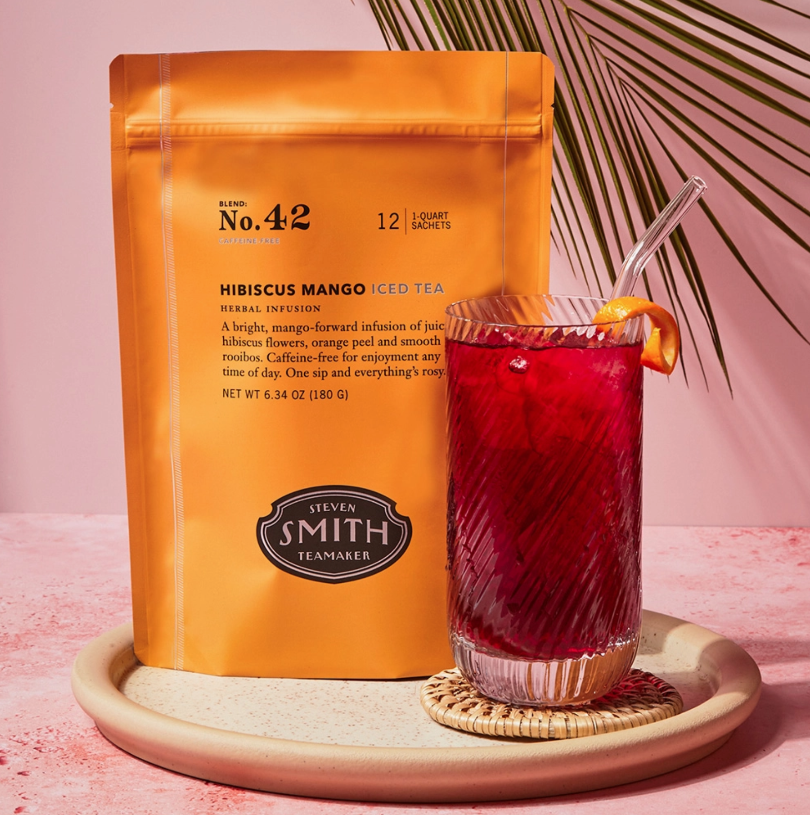 A bright orange Faire Smith Tea bag of Hibiscus Mango Iced Tea blend No. 42 sits next to a clear glass of caffeine-free iced tea, garnished with an orange slice, on a textured pink surface. Perfectly refreshing!.