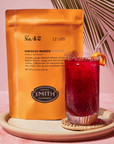 A bright orange Faire Smith Tea bag of Hibiscus Mango Iced Tea blend No. 42 sits next to a clear glass of caffeine-free iced tea, garnished with an orange slice, on a textured pink surface. Perfectly refreshing!.