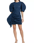 A person in a navy Pia Mini Dress by Rhode, featuring puffed short sleeves, a ruched asymmetric skirt with delicate embellishments, elegantly draped on one side, paired with transparent heels.