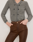 A person wearing The Court Cardi by FRAME, featuring black and white stripes, contrast stripe trim, black buttons, and a V neckline, stands against a plain white background with their hands in their pockets, paired with dark brown pants.