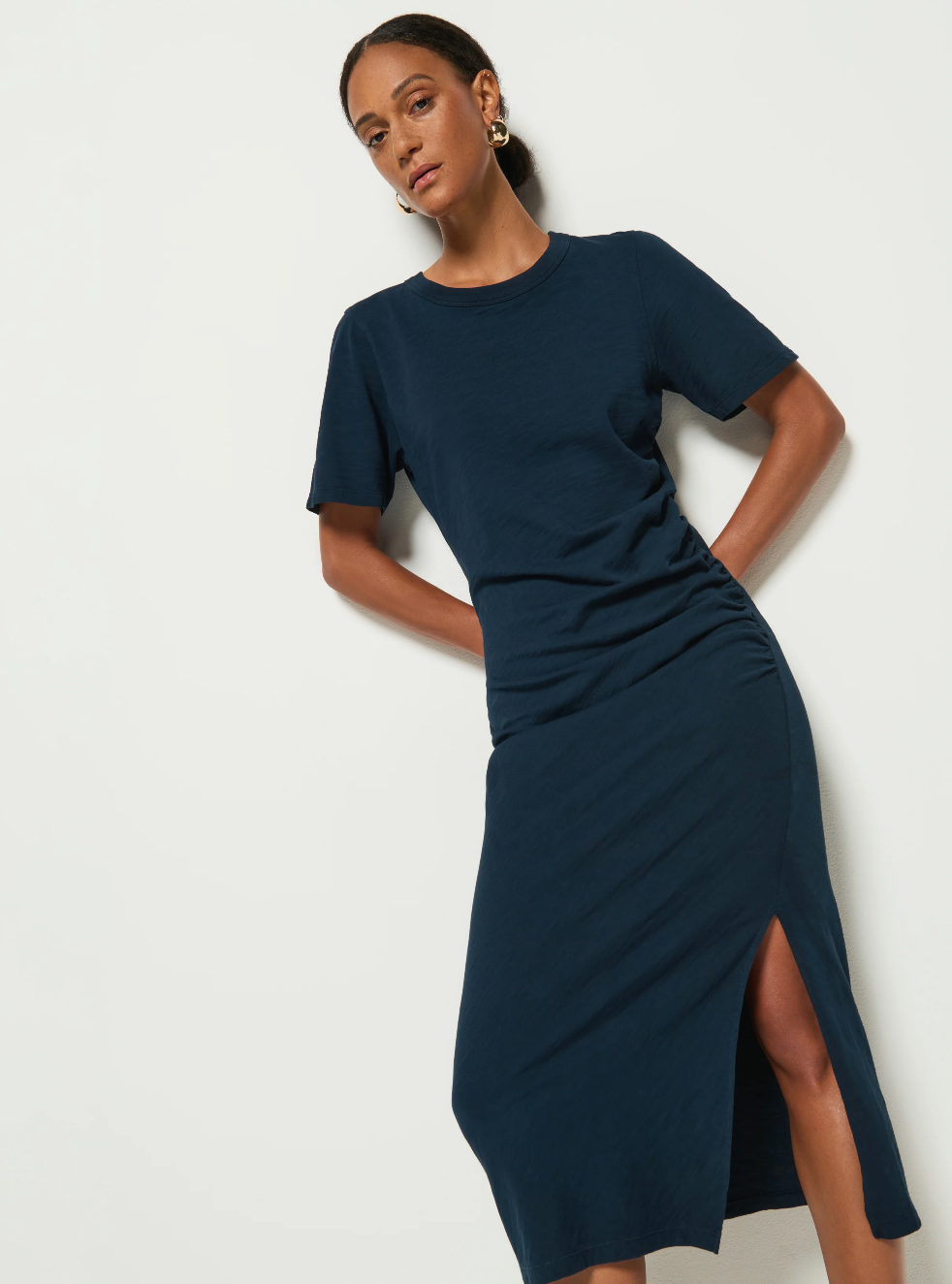 A person in a navy blue Cole T-shirt Dress by Nation LTD, reminiscent of the Night Sky, stands against a white backdrop. The dress features a side slit. They have short dark hair and wear gold hoop earrings. Their pose is relaxed, hands resting gently on their lower back.