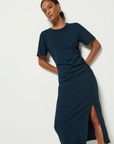 A person in a navy blue Cole T-shirt Dress by Nation LTD, reminiscent of the Night Sky, stands against a white backdrop. The dress features a side slit. They have short dark hair and wear gold hoop earrings. Their pose is relaxed, hands resting gently on their lower back.