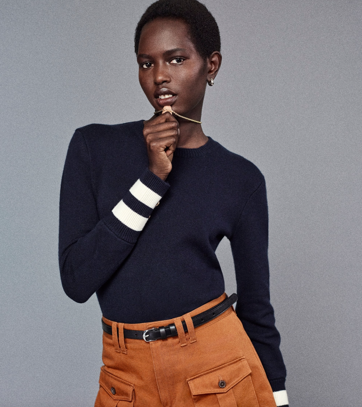 A person poses against a gray background, wearing FRAME's navy sweater with white striped cuffs and The Scout Short. They hold a necklace near their lips while looking at the camera, blending casual style with elegance.