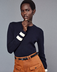 A person poses against a gray background, wearing FRAME's navy sweater with white striped cuffs and The Scout Short. They hold a necklace near their lips while looking at the camera, blending casual style with elegance.