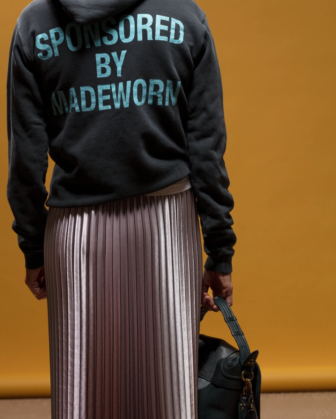 Against an orange backdrop, a person models the MadeWorn Artist Hoodie by Made Worn in heavyweight French terry cotton with "SPONSORED BY MADE WORN" on the back, paired with a pleated lavender skirt and accessorized with a black bag. Handmade in Los Angeles.