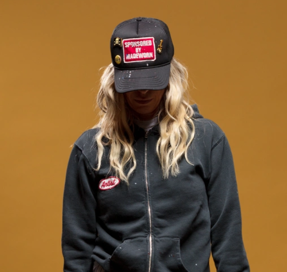 A person with long wavy hair wears a black hoodie and the limited edition MadeWorn Sponsor Hat by Made Worn, featuring a "Sponsored by Madeworn" patch. The adjustable back strap is visible as they face forward against an ochre background, their face mostly hidden by the cap.