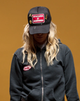 A person with long wavy hair wears a black hoodie and the limited edition MadeWorn Sponsor Hat by Made Worn, featuring a "Sponsored by Madeworn" patch. The adjustable back strap is visible as they face forward against an ochre background, their face mostly hidden by the cap.