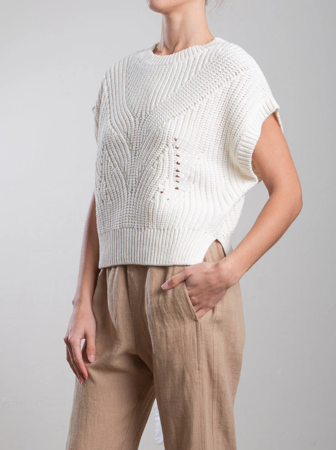 A person in a Pharaoh Edie Shell, notable for its ribbed accents and intricate design, pairs it with light beige high-waisted pants. With one hand in their pocket, they stand against a plain light gray background.