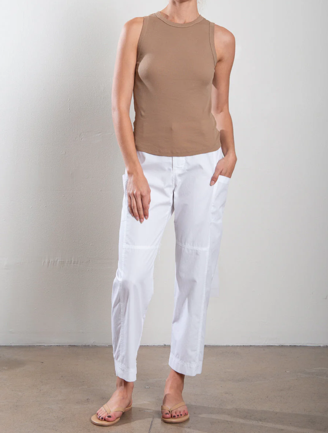 Someone stands against a plain wall wearing a sleeveless beige top and Pharaoh's Torrance Pant, featuring stylish barrel-leg design and an elasticized waist. They casually have their hands in their pockets and sport flip-flops.