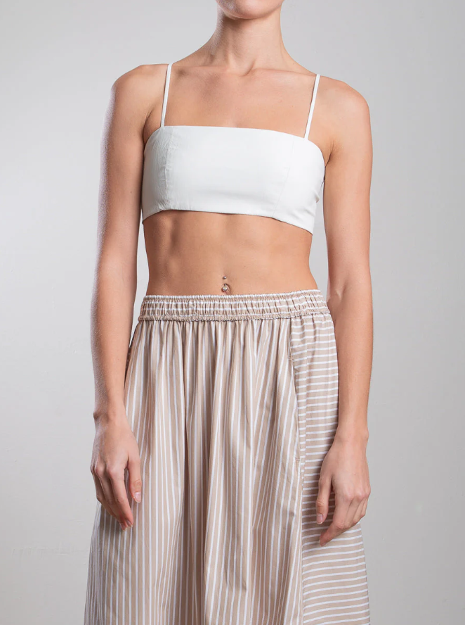 A person showcases the Pharaoh Toga Bralette, a white spaghetti strap piece, paired with a beige skirt featuring white vertical stripes, set against a plain gray backdrop.