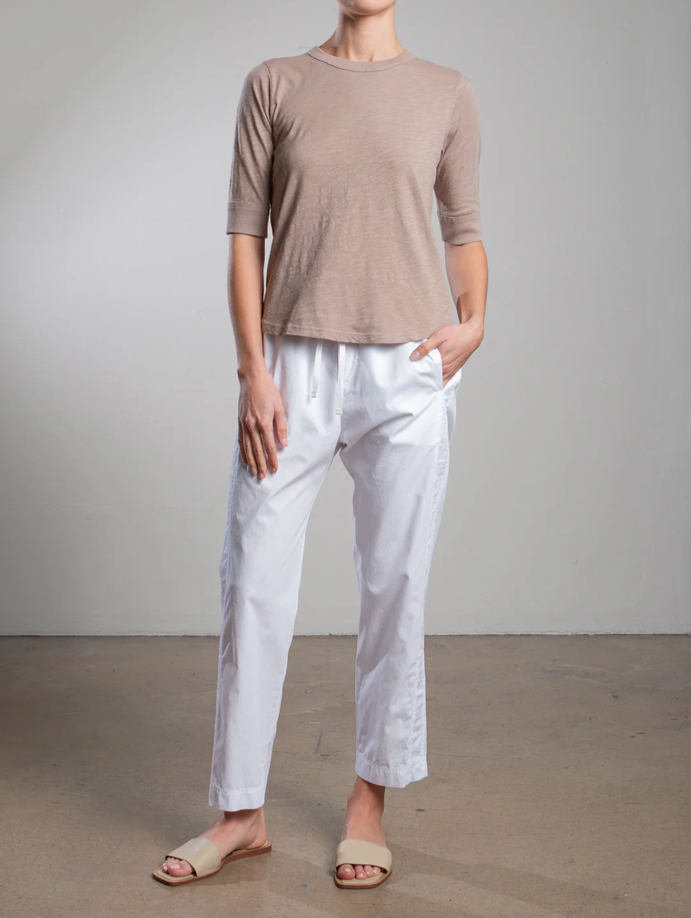 Against a plain backdrop, a person exudes casual style in the Andy Pant by Pharaoh, paired with a light brown short-sleeve top featuring sateen accents. Their ensemble is completed with relaxed-fit white pants and beige slip-on sandals, while one hand rests in their pocket.