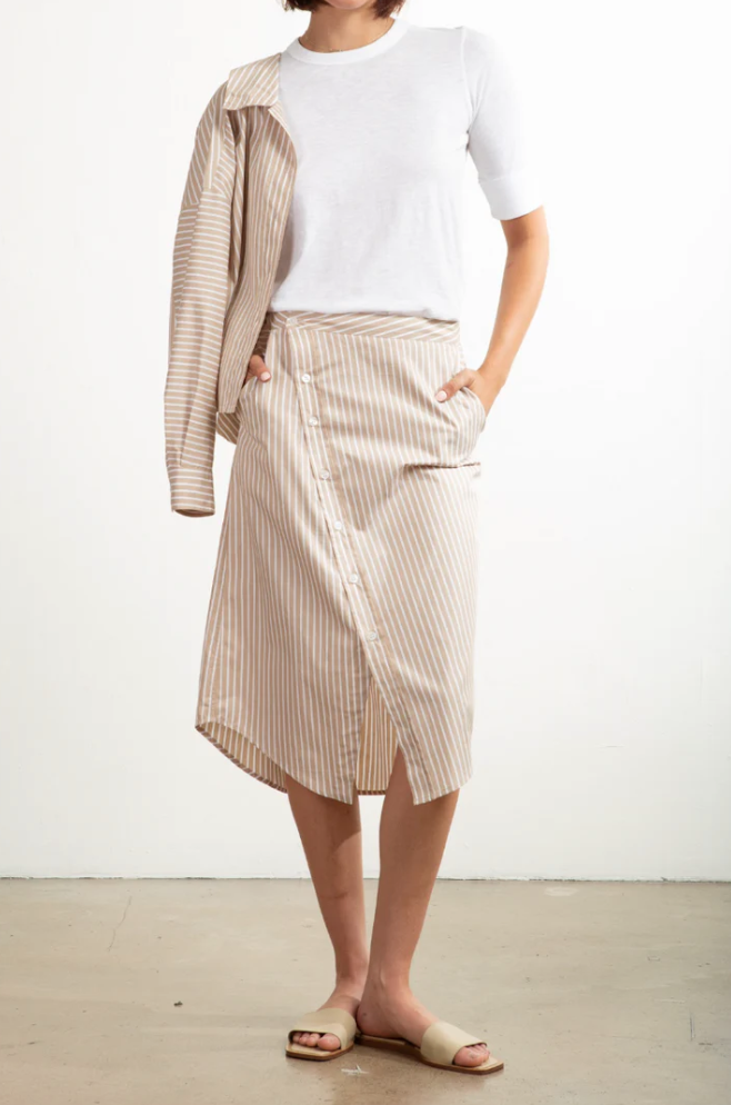 A person in a white t-shirt has tucked it into Pharaoh's Nico Skirt, featuring beige and white vertical stripes, an asymmetric placket, and front slash pockets. Draped over one shoulder is the matching striped shirt. They wear light-colored sandals while standing on a plain surface.