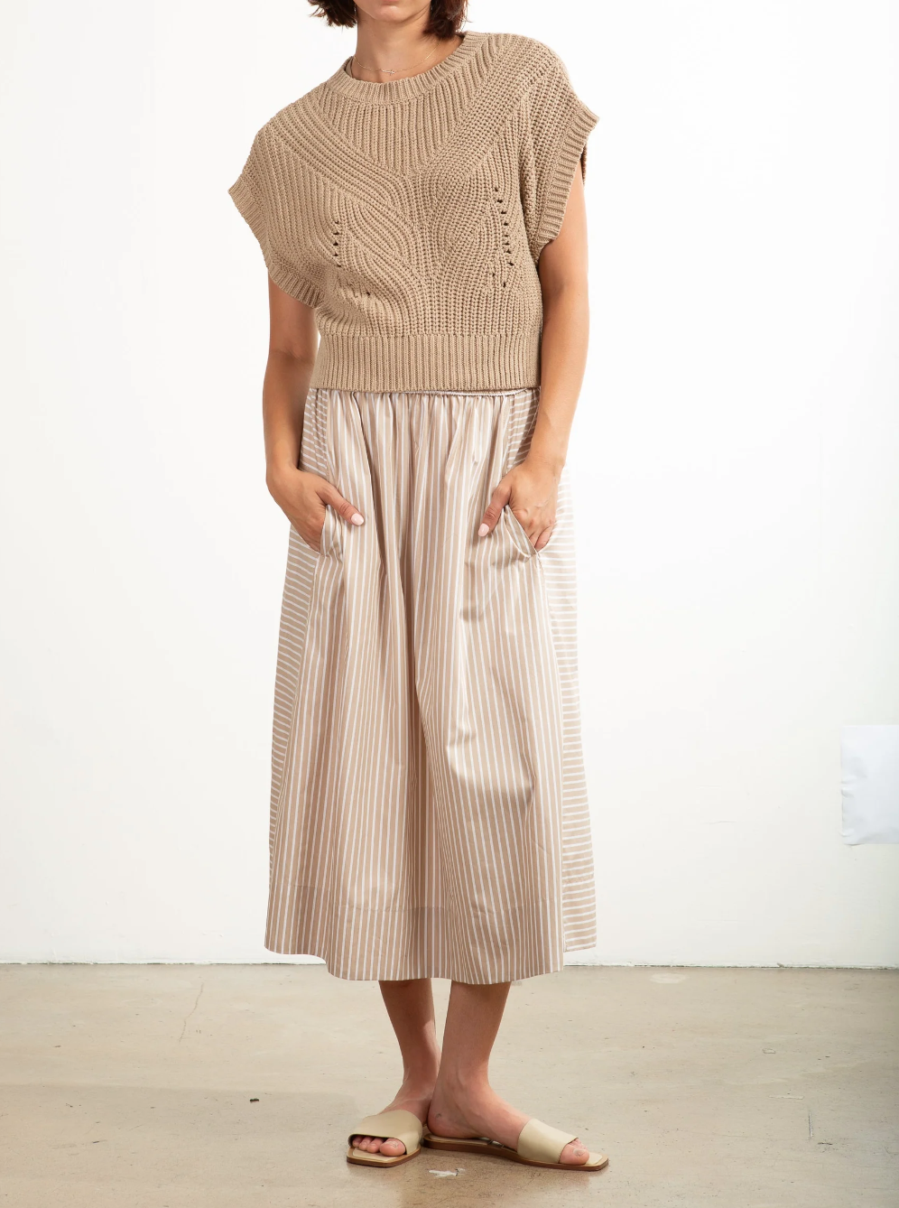 A person in a beige, short-sleeved knit top and Pharaoh's Claudia Skirt, a high-waisted 100% cotton midi with side panel details, stands hands-in-pockets barefoot on concrete against a white backdrop.