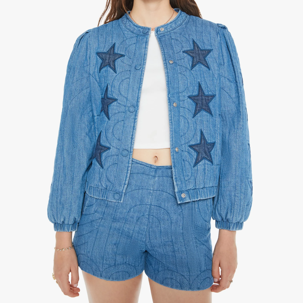 A woman in a blue denim jacket by Mother called "The Rough Puff," featuring puff shoulders, blue star patches, a button-up front, and elasticized cuffs. She's wearing a white crop top and shorts with long brown hair cascading down against a plain white background.