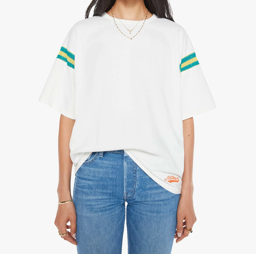 A woman wears "The Sleep Over Inset" by Mother, an oversized white t-shirt with green and yellow striped sleeves, tucked into blue jeans. Her long hair is complemented by gold necklaces and rings. The shirt features an orange logo near the hem, ideal for a casual vibe.